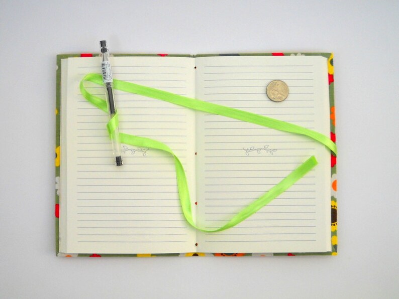 May include: A green ribbon is tied around a closed notebook with a floral pattern on the cover. A pen and a coin are on the open pages.