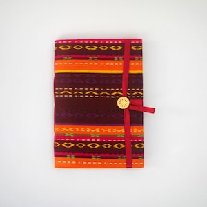 K&ouml;nnte beinhalten: Ein farbenfrohes Notizbuch mit roter Schleifenverschlusses und einem Holz Knopf. Der Einband zeigt ein geometrisches Muster in Orange-, Rot-, Lila- und Gelbt&ouml;nen.