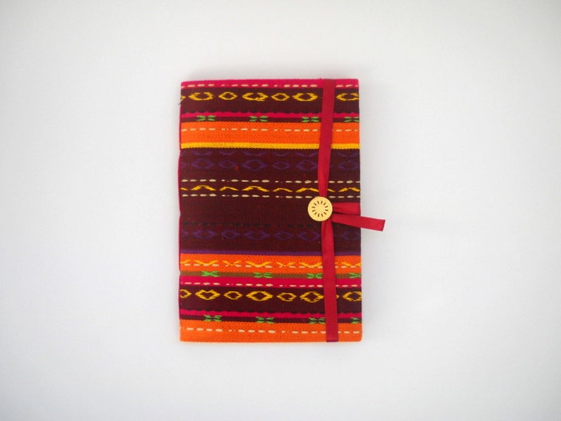 K&ouml;nnte beinhalten: Ein farbenfrohes Notizbuch mit roter Schleifenverschlusses und einem Holz Knopf. Der Einband zeigt ein geometrisches Muster in Orange-, Rot-, Lila- und Gelbt&ouml;nen.