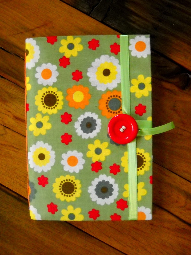 May include: A green fabric notebook with a floral pattern in shades of white, yellow, orange, and red. The notebook has a green ribbon closure and a red button.