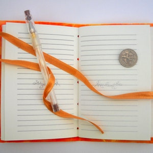 May include: An orange notebook with lined pages, a clear pen, and a gold coin. The notebook is tied closed with an orange ribbon.