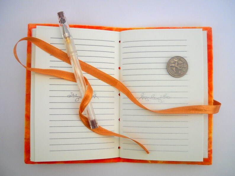 May include: An orange notebook with lined pages, a clear pen, and a gold coin. The notebook is tied closed with an orange ribbon.