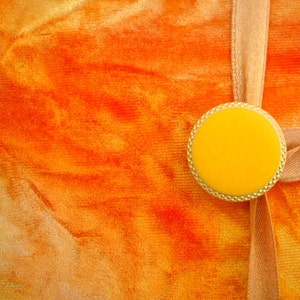 May include: A close-up of a bright orange fabric background with a gold-colored circular embellishment with a yellow center. The embellishment is attached to a light brown ribbon.