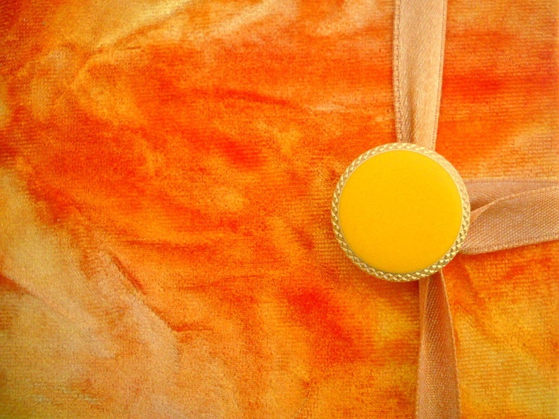 May include: A close-up of a bright orange fabric background with a gold-colored circular embellishment with a yellow center. The embellishment is attached to a light brown ribbon.