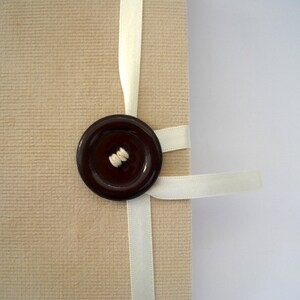 May include: A beige fabric journal with a white ribbon tied around it. A brown button with a white thread detail is attached to the ribbon.