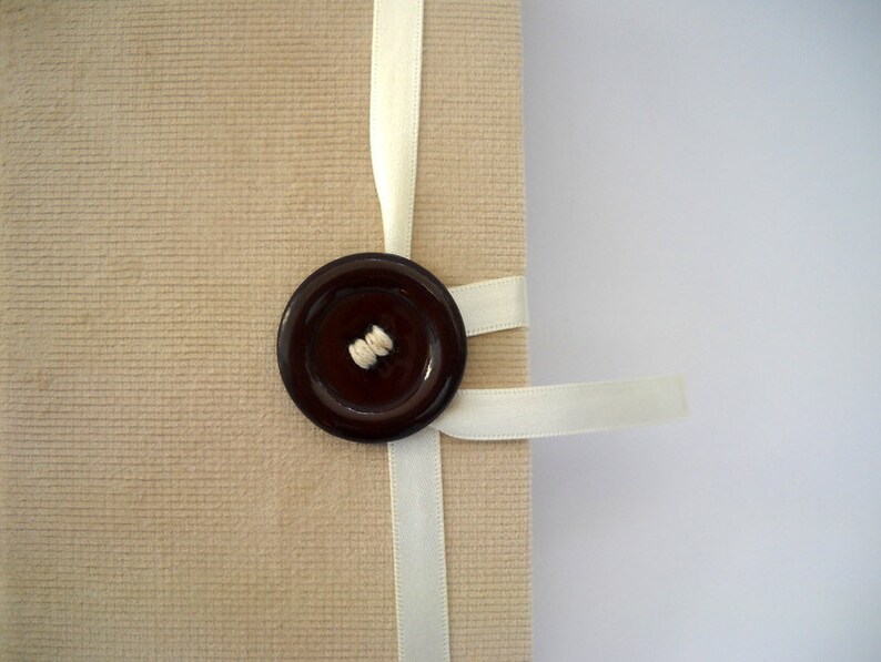 May include: A beige fabric journal with a white ribbon tied around it. A brown button with a white thread detail is attached to the ribbon.