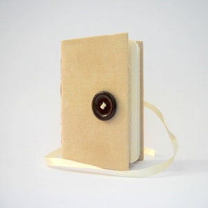May include: A small, beige linen-covered journal with a brown button closure and a cream-colored ribbon bookmark.
