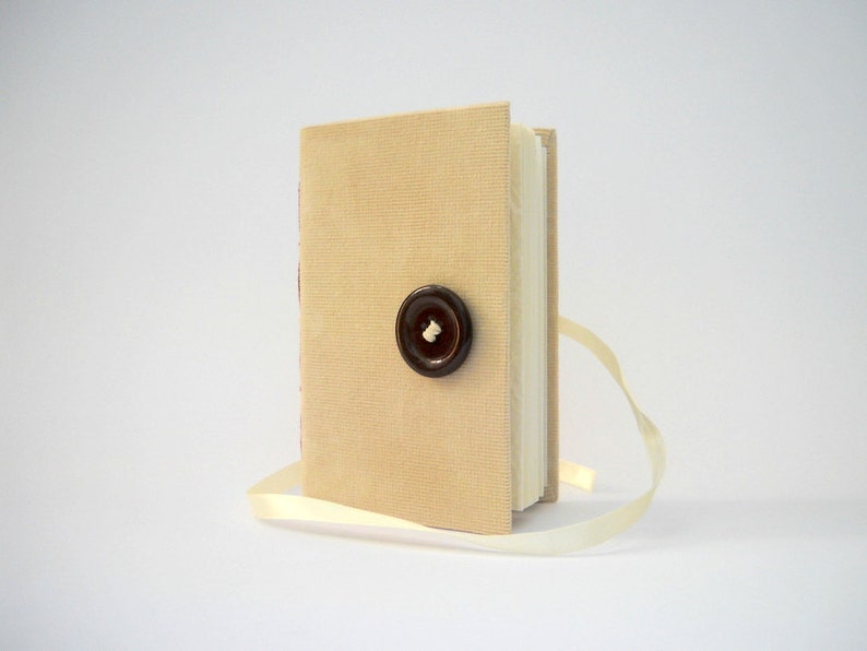 May include: A small, beige linen-covered journal with a brown button closure and a cream-colored ribbon bookmark.