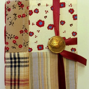 May include: A handmade fabric-covered journal with a red ribbon closure and a vintage coin embellishment. The cover is made of patchwork fabric in various patterns, including plaid, floral, and polka dots.