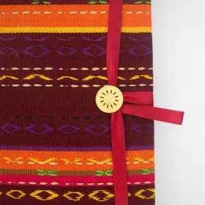 K&ouml;nnte beinhalten: Ein rotes Band um ein burgunderfarbenes Stofftagebuch mit einem goldenen Zierknopf. Der Stoff hat ein buntes gewebtes Muster.