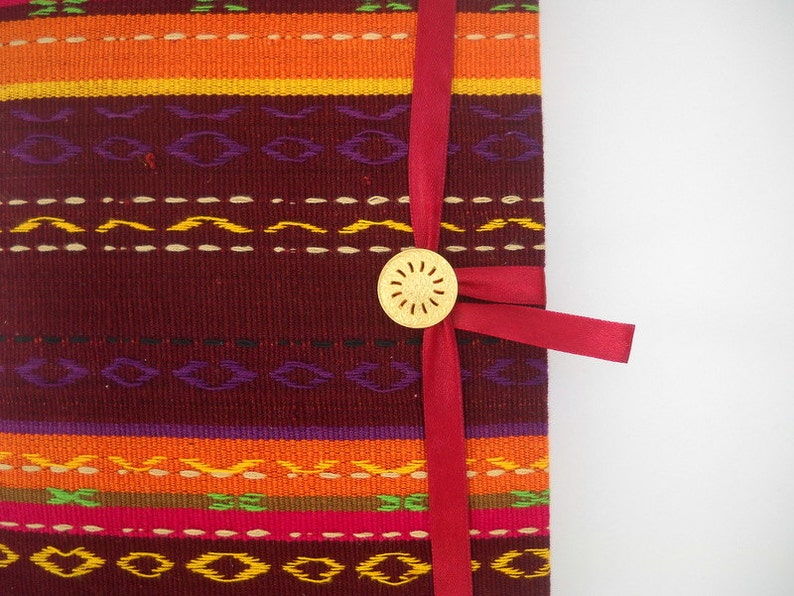 K&ouml;nnte beinhalten: Ein rotes Band um ein burgunderfarbenes Stofftagebuch mit einem goldenen Zierknopf. Der Stoff hat ein buntes gewebtes Muster.