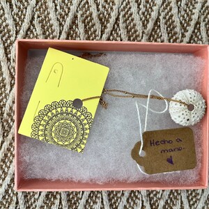 May include: A pink box with a white interior contains a gold necklace with a white sea urchin pendant. The box also includes two yellow cards with black designs and a brown card with the text "Hecho a mano" in purple.