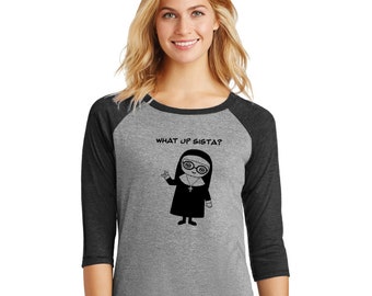 May include: A gray and black baseball tee shirt with a cartoon nun wearing glasses and a cross. The text "WHAT UP SISTA?" is printed on the shirt.