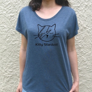May include: A blue t-shirt with a black graphic of a cat with a lightning bolt across its eye. The text "Kitty Stardust" is printed below the cat.