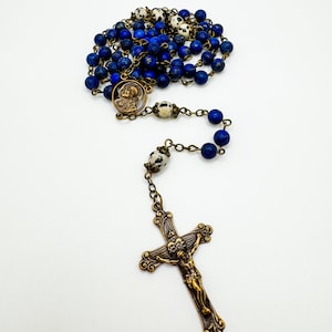 May include: A rosary with deep blue beads and a bronze crucifix. The beads are interspersed with white and black speckled beads. The crucifix features a detailed depiction of Jesus Christ.