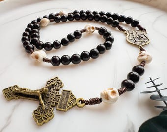 May include: A rosary with black beads and skull-shaped accents. The crucifix and connecting pieces are bronze-colored. The rosary is displayed on a white surface.