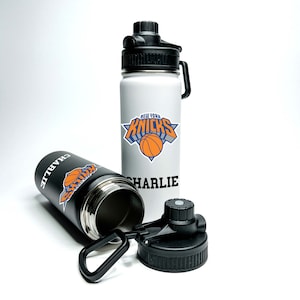 Puede incluir: Dos botellas de agua de acero inoxidable con tapas negras y un logotipo de los New York Knicks en blanco y negro. Una botella es blanca con el logotipo y el nombre "CHARLIE" impreso en ella. La otra botella es negra con el nombre "CHARLIE" impreso en ella.