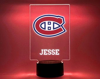 May include: A red, white, and blue Montreal Canadiens logo with the name "JESSE" printed below it on a clear acrylic plaque. The plaque is lit from behind and sits on a black base.