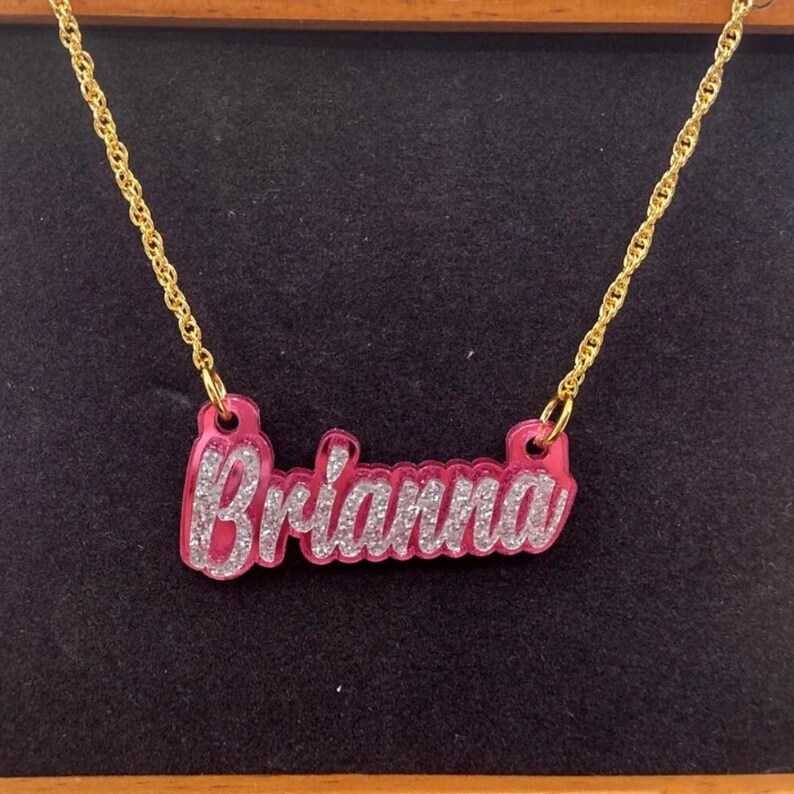 K&ouml;nnte beinhalten: Eine goldene Kettenkette mit einem rosafarbenen Namensschild-Anh&auml;nger, auf dem "Brianna" in silbernen Glitzerbuchstaben steht.
