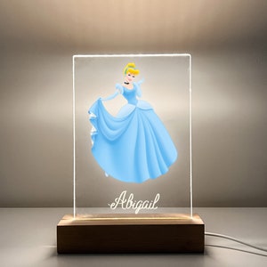 K&ouml;nnte beinhalten: Eine durchsichtige Acryl-Leuchtanzeige mit einem Cartoon-Bild von Aschenputtel in einem blauen Kleid. Der Sockel ist aus Holz und der Name "Abigail" ist auf dem Display aufgedruckt.