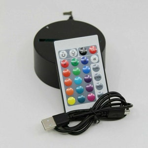 K&ouml;nnte beinhalten: Eine schwarze runde Basis mit einem USB-Kabel und einer wei&szlig;en Fernbedienung mit 16 Tasten. Die Fernbedienung hat Tasten zum Ein- und Ausschalten, Blinken, Stroboskop, Verblassen, Gl&auml;tten und verschiedene Farben.