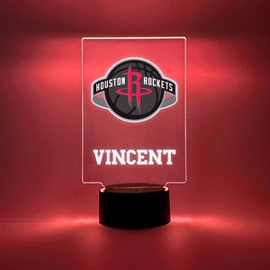 Puede incluir: Una luz nocturna de acrílico transparente con una base negra. La luz presenta un logotipo de los Houston Rockets con el texto "HOUSTON ROCKETS" y el nombre "VINCENT" debajo del logotipo.