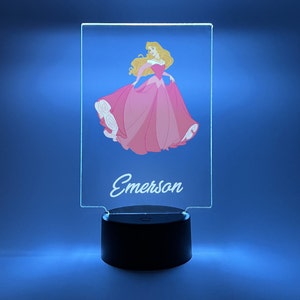 May include: A clear acrylic night light with a pink princess illustration and the name "Emerson" printed on the front. The night light has a black base.
