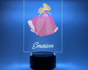 Könnte beinhalten: Ein klares Acryl-Nachtlicht mit einer rosa Prinzessinnen-Illustration und dem Namen "Emerson" auf der Vorderseite. Das Nachtlicht hat einen schwarzen Sockel.