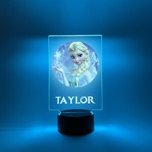 K&ouml;nnte beinhalten: Ein blaues und wei&szlig;es Nachtlicht mit einer durchsichtigen Acrylplatte, die ein Bild von Elsa aus Frozen zeigt. Die Platte wird von hinten beleuchtet, wodurch ein leuchtender Effekt entsteht. Der Name "TAYLOR" ist unter dem Bild aufgedruckt.