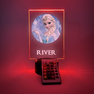 K&ouml;nnte beinhalten: Ein personalisiertes LED-Nachtlicht mit einem Bild von Elsa aus Frozen. Das Licht steht auf einem schwarzen Sockel mit einer Fernbedienung. Der Name "RIVER" ist auf das Licht gedruckt.