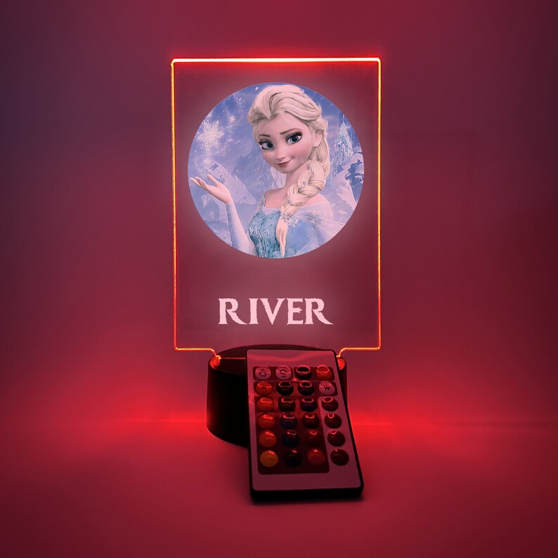 K&ouml;nnte beinhalten: Ein personalisiertes LED-Nachtlicht mit einem Bild von Elsa aus Frozen. Das Licht steht auf einem schwarzen Sockel mit einer Fernbedienung. Der Name "RIVER" ist auf das Licht gedruckt.