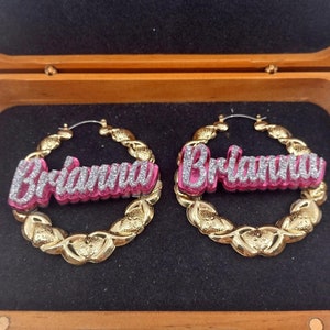 K&ouml;nnte beinhalten: Goldene Creolen mit dem Namen "Brianna" in pink und silbernem Glitzer.