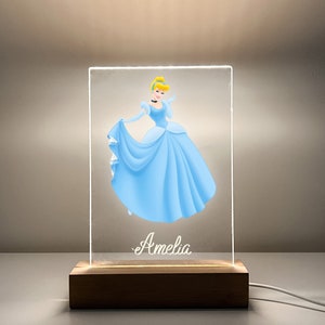 K&ouml;nnte beinhalten: Ein klares Acryl-Nachtlicht mit einer Cinderella-Illustration und dem Namen "Amelia" auf der Basis. Das Licht steht auf einem Holzsockel mit einem Kabel.