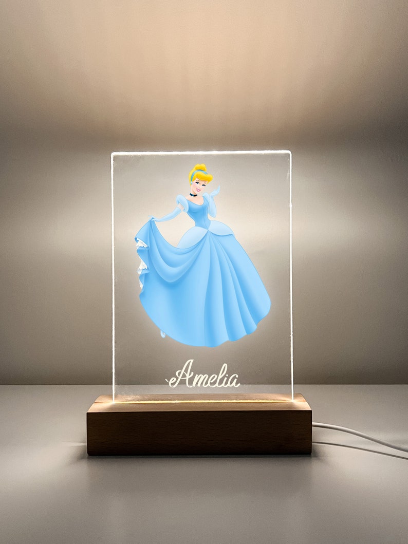K&ouml;nnte beinhalten: Ein klares Acryl-Nachtlicht mit einer Cinderella-Illustration und dem Namen "Amelia" auf der Basis. Das Licht steht auf einem Holzsockel mit einem Kabel.