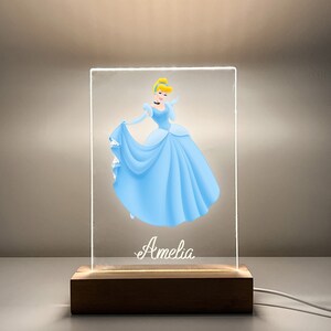 K&ouml;nnte beinhalten: Ein klares Acryl-Nachtlicht mit einer Cinderella-Illustration und dem Namen "Amelia" auf der Basis. Das Licht steht auf einem Holzsockel mit einem Kabel.
