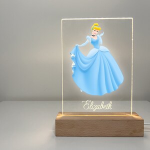 K&ouml;nnte beinhalten: Ein leuchtendes Acrylschild mit einem Cartoon-Bild von Aschenputtel in einem blauen Kleid. Das Schild hat einen Holzsockel und der Name "Elizabeth" ist unten in Kursivschrift geschrieben.