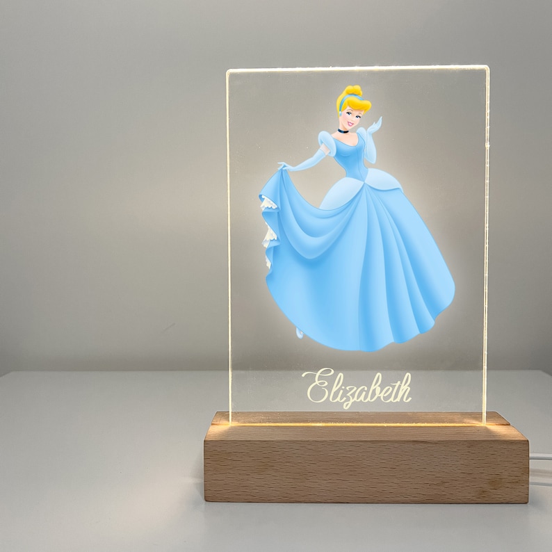 K&ouml;nnte beinhalten: Ein leuchtendes Acrylschild mit einem Cartoon-Bild von Aschenputtel in einem blauen Kleid. Das Schild hat einen Holzsockel und der Name "Elizabeth" ist unten in Kursivschrift geschrieben.