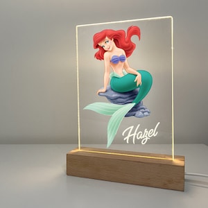 Puede incluir: Un letrero acrílico iluminado con una imagen de dibujos animados de Ariel, la Sirenita, con una cola verde y un sujetador de concha de mar púrpura. El letrero tiene una base de madera y el nombre "Hazel" está escrito en blanco debajo de la imagen.