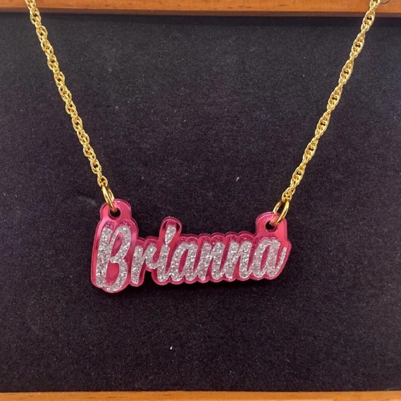K&ouml;nnte beinhalten: Eine goldene Kettenkette mit einem rosafarbenen Namensschild-Anh&auml;nger, auf dem "Brianna" in silbernen Glitzerbuchstaben steht.