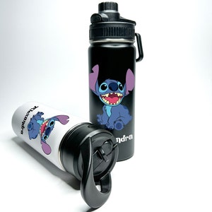Puede incluir: Dos botellas de agua de acero inoxidable en blanco y negro con un personaje de dibujos animados azul en cada una. El personaje es Stitch de la película de Disney Lilo & Stitch. Las botellas tienen tapas negras con una asa de transporte. El texto "Alexandra" y "Sandra" está impreso en las botellas.