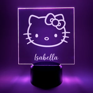 May include: A white acrylic night light with a Hello Kitty design and the name "Isabella" engraved on the bottom. The light is on a black base and is illuminating a purple surface.