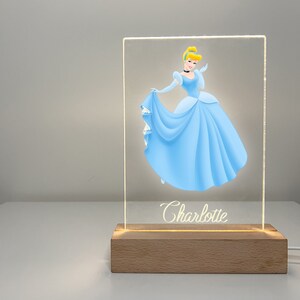 K&ouml;nnte beinhalten: Ein klares Acryl-Nachtlicht mit einer Disney-Cinderella-Illustration und dem Namen "Charlotte" auf der Unterseite. Das Licht steht auf einem Holzsockel.