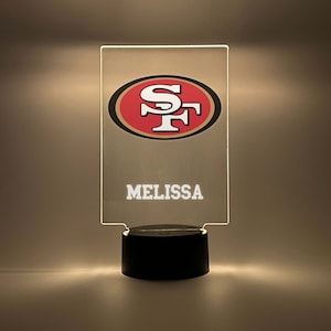 Puede incluir: Una luz nocturna de acrílico transparente con un logotipo rojo y blanco de los San Francisco 49ers y el nombre "MELISSA" impreso en la parte delantera. La luz está sobre una base negra.