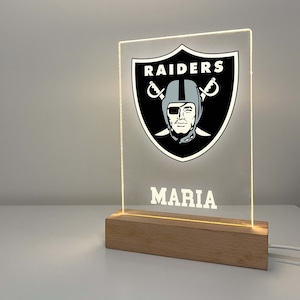 May include: A clear acrylic Raiders team logo night light with a wooden base. The Raiders logo is in black, white, and grey. The name "MARIA" is printed below the logo. The light is illuminated.