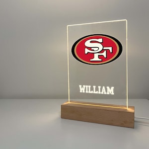 Puede incluir: Un letrero de acrílico transparente con un logotipo de los San Francisco 49ers en rojo y dorado y el nombre "William" en texto blanco. El letrero está iluminado desde atrás y se asienta sobre una base de madera.
