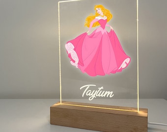 Könnte beinhalten: Ein rosa-weißes Acryl-Nachtlicht mit einer Prinzessin in einem rosa Kleid. Der Name "Taytum" ist in Weiß unter der Prinzessin geschrieben. Das Nachtlicht hat einen Holzsockel.