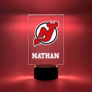 May include: A clear acrylic night light with a red and white New Jersey Devils logo and the name "NATHAN" printed below. The light is on a black base.