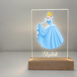 K&ouml;nnte beinhalten: Ein leuchtendes Acrylschild mit einem Cartoon-Bild von Aschenputtel in einem blauen Kleid. Das Schild hat einen Holzsockel und der Name "Elizabeth" ist unten in Kursivschrift geschrieben.