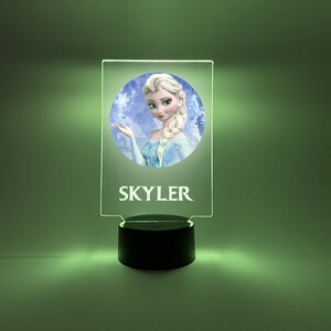 K&ouml;nnte beinhalten: Ein klares Acryl-Nachtlicht mit einem runden Bild einer Comic-Prinzessin mit blonden Haaren und einem blauen Kleid. Das Bild ist von einem wei&szlig;en Rand umgeben und der Name "SKYLER" ist unter dem Bild gedruckt.