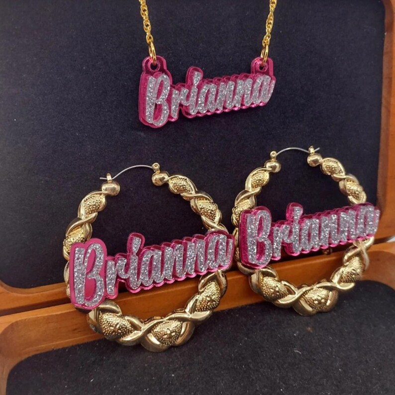 K&ouml;nnte beinhalten: Eine goldene Kettenkette und Creolen mit einem rosafarbenen Namensschild-Anh&auml;nger, auf dem "Brianna" in silbernen Glitzerbuchstaben steht.